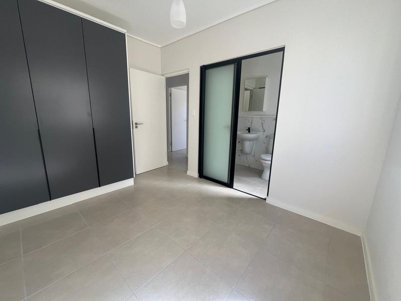 A modern, empty bedroom featuring sleek dark built-in cupboards, neutral tiled flooring, and a sliding door leading to an en-suite bathroom.