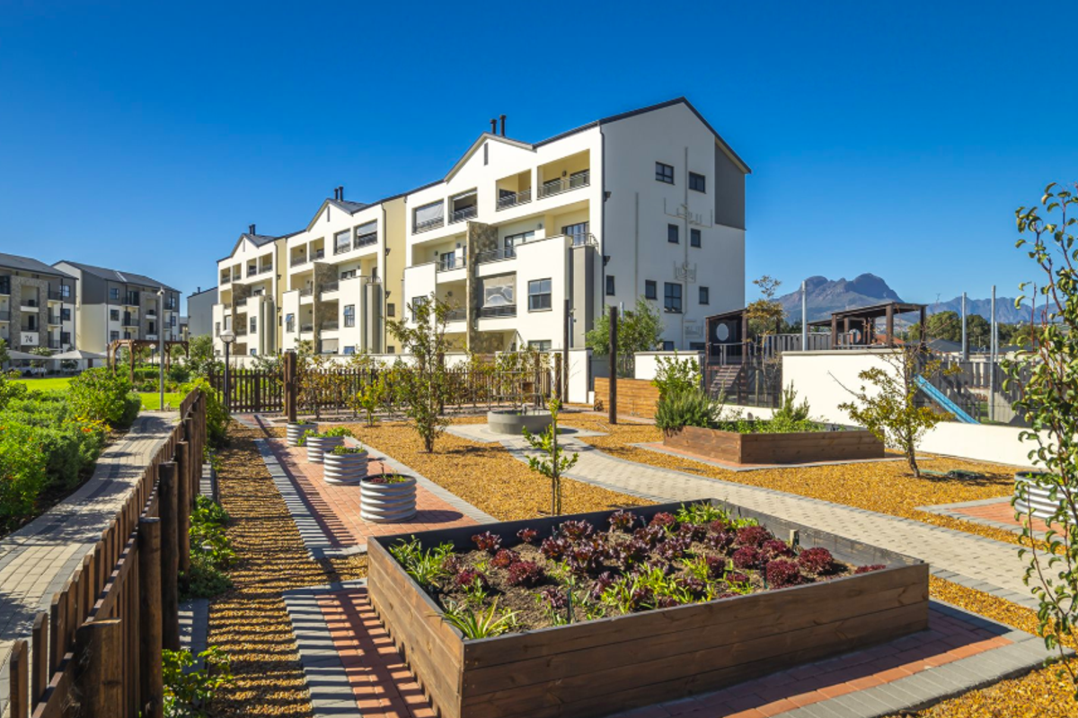 A well-maintained communal garden area featuring raised timber planter beds, paved pathways, and modern apartment buildings in the background.