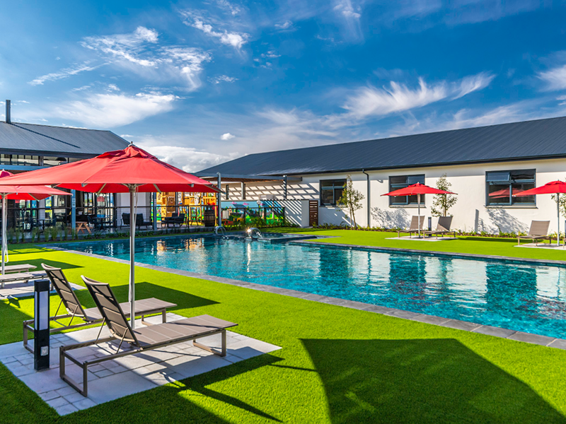 A well-maintained communal swimming pool area featuring artificial turf, modern pool surrounds, and clean building exteriors under a clear blue sky.