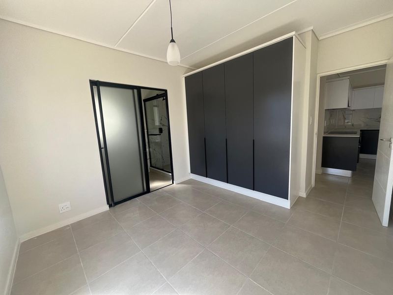 A modern, empty bedroom featuring sleek dark built-in cupboards, neutral tiled flooring, and a contemporary frosted glass door leading to an en-suite bathroom.