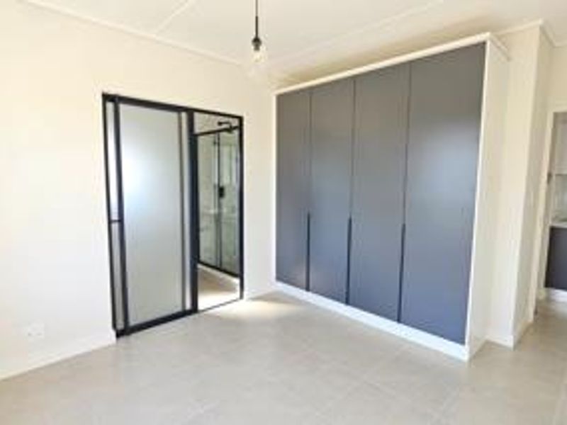 A modern, bright bedroom featuring sleek grey built-in cupboards, neutral tiled flooring, and a contemporary glass-panelled door leading to an en-suite bathroom.