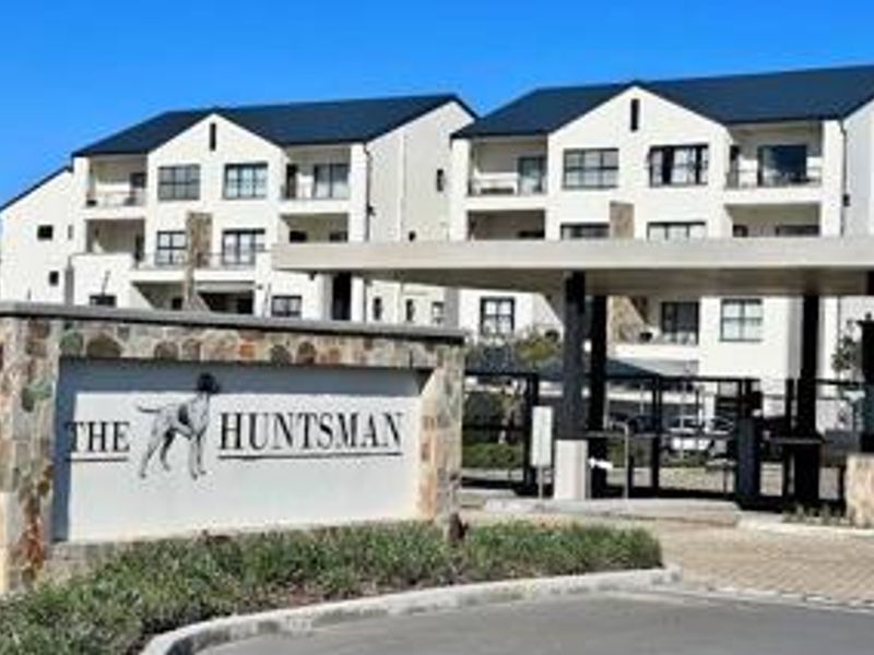 Front entrance view of The Huntsman apartment complex featuring a stone-clad security gate, paved driveway, and modern multi-story residential buildings.