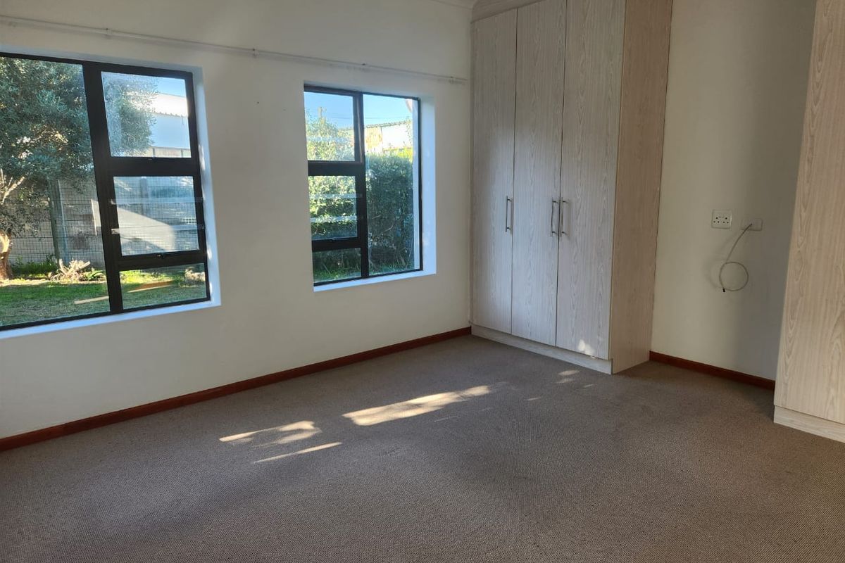 A bright, empty bedroom featuring neutral carpeting, built-in wooden cupboards, and large windows overlooking the garden.