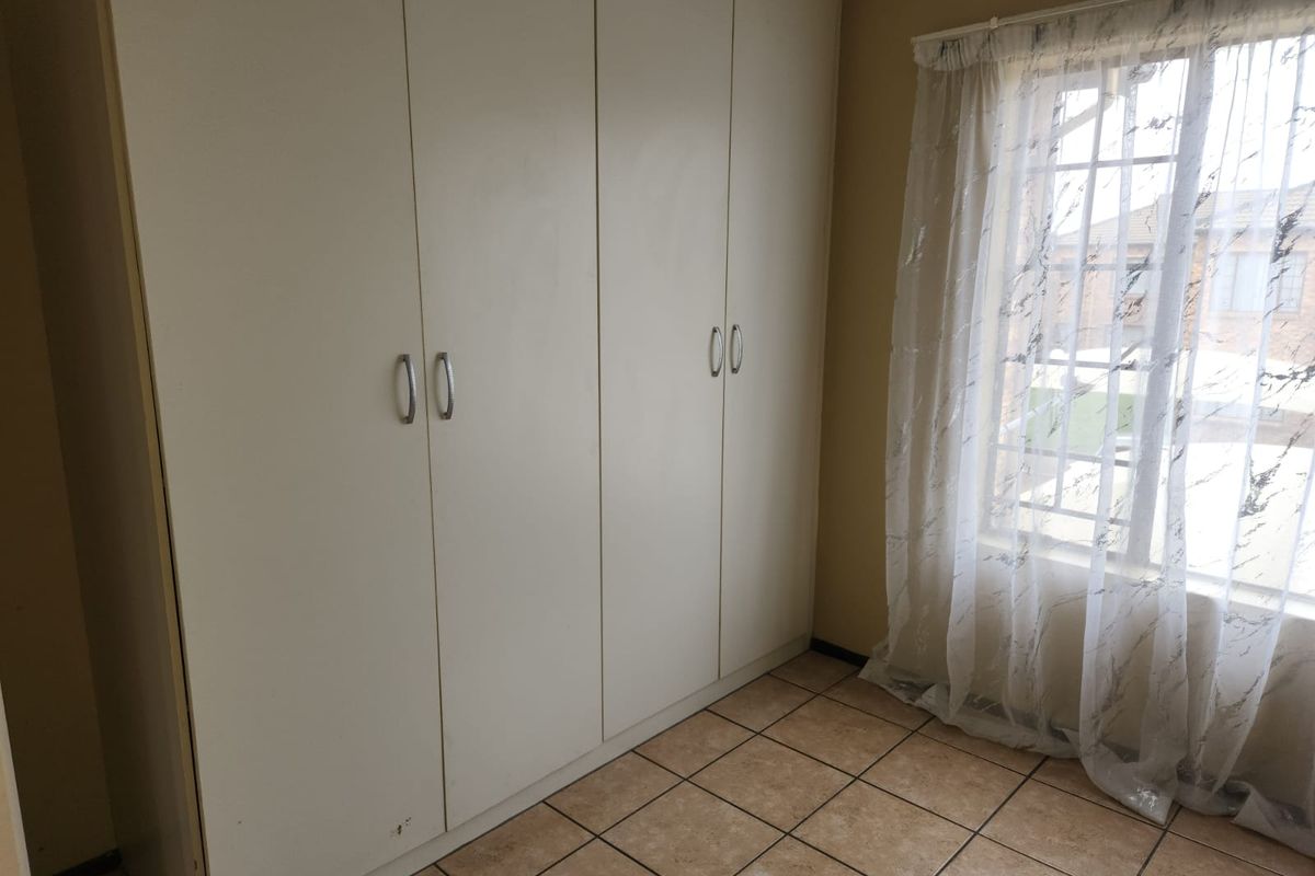 A bedroom featuring built-in cupboards, tiled flooring, and a window with security bars, offering a functional and neutral space.