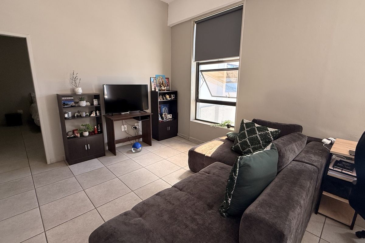 A living room featuring neutral wall tones, tiled flooring, and a large window with a roller blind, providing natural light to the space.