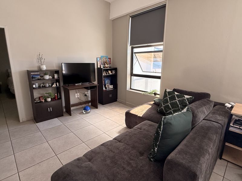 A living room featuring neutral wall tones, tiled flooring, and a large window with a roller blind, providing natural light to the space.