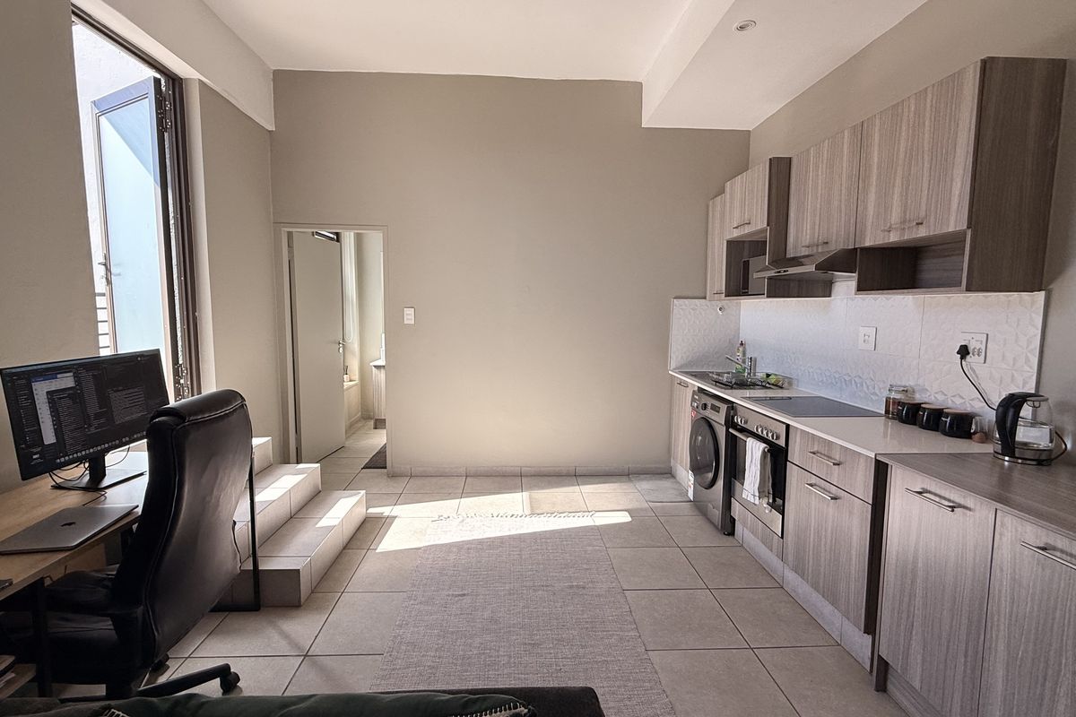 Open-plan living area featuring a modern kitchen with wood-finish cabinetry, integrated appliances, and tiled flooring, leading to an adjacent workspace.