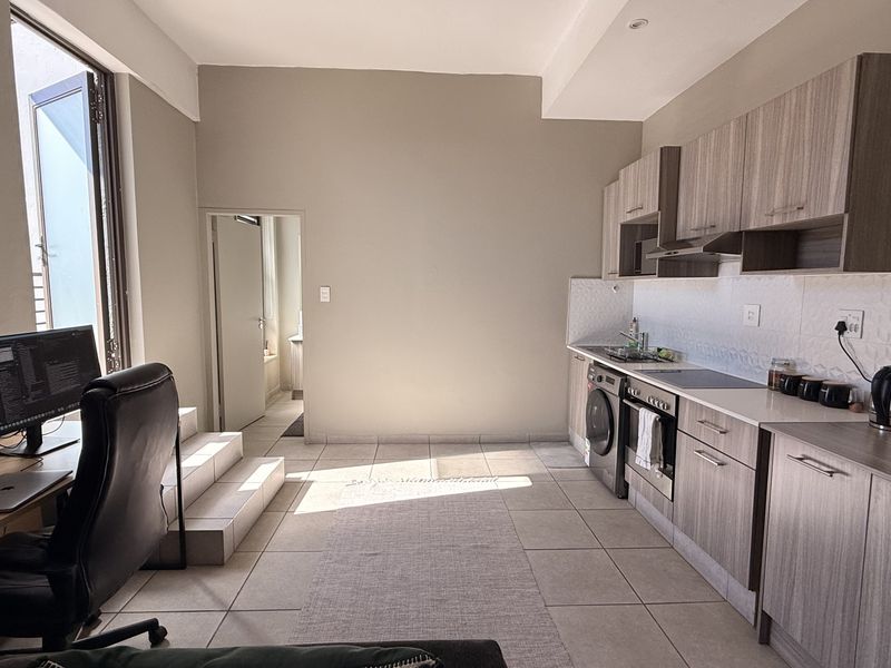 Open-plan living area featuring a modern kitchen with wood-finish cabinetry, integrated appliances, and tiled flooring, leading to an adjacent workspace.