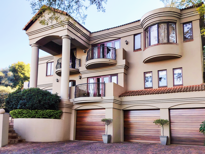 A grand multi-story residence featuring a triple garage with timber doors, elegant balconies with wrought iron railings, and classic architectural columns.