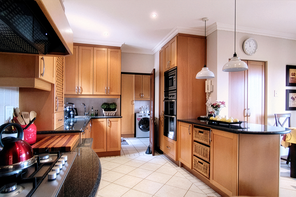 A well-appointed kitchen featuring light wood cabinetry, granite countertops, built-in appliances, and an adjoining scullery area with tiled flooring throughout.