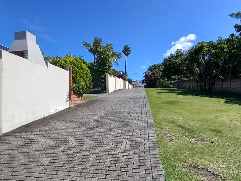 A paved driveway leading towards the property, featuring a boundary wall, a built-in braai area, and a well-maintained lawn.