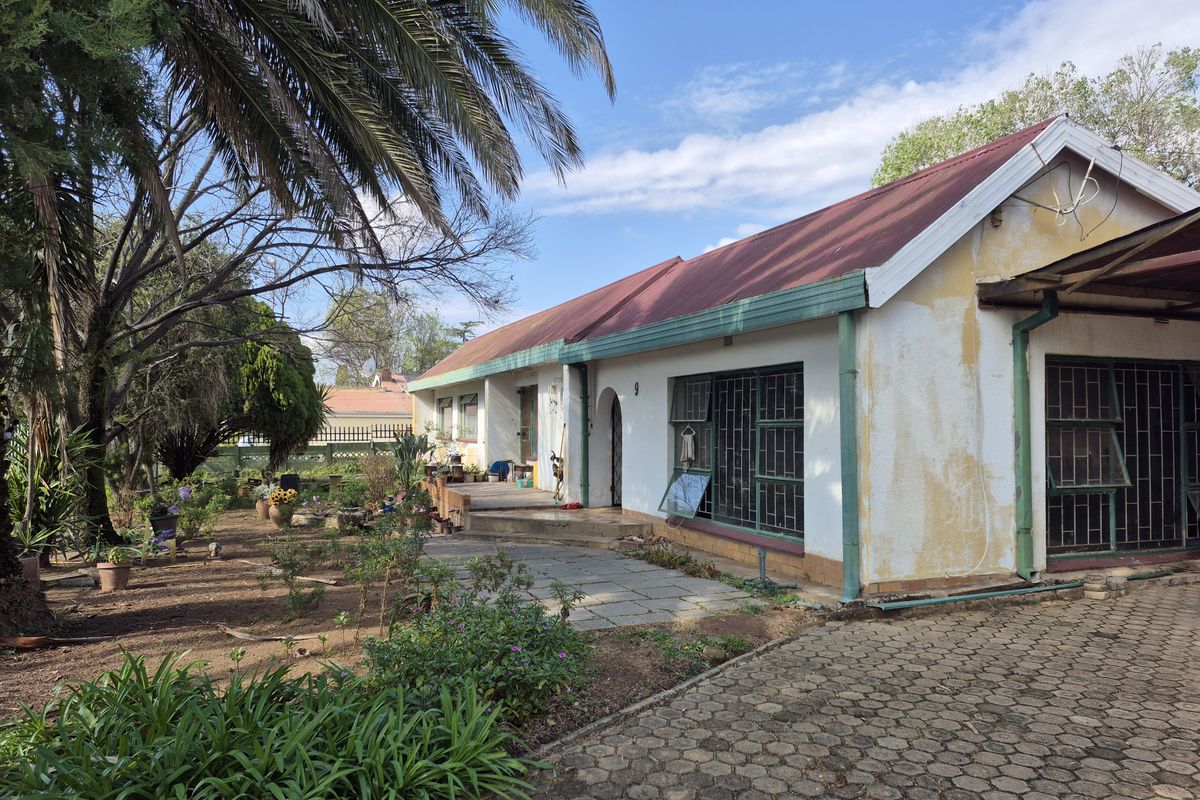 Rear view of the property featuring a paved driveway, stone patio area, and metal roof, showing signs of exterior wear and maintenance requirements.
