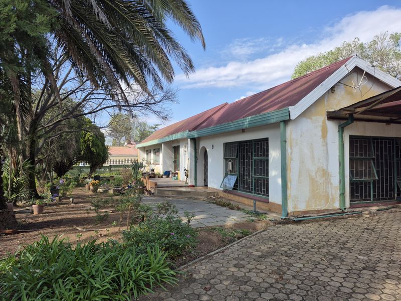 Rear view of the property featuring a paved driveway, stone patio area, and metal roof, showing signs of exterior wear and maintenance requirements.