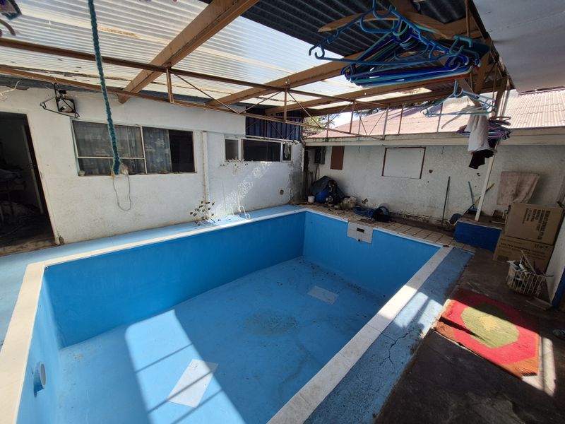 An outdoor swimming pool area featuring a covered structure with a corrugated roof, showing signs of wear and requiring maintenance to the pool and finishes.