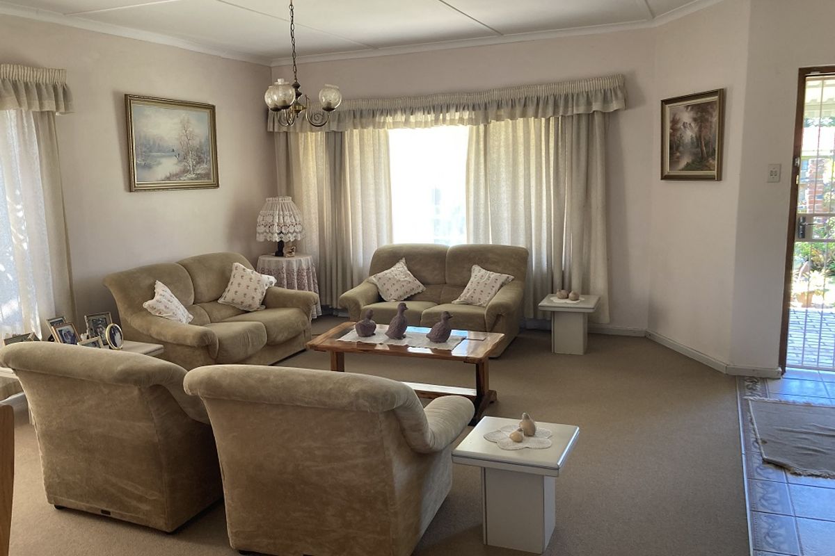 A spacious living room featuring neutral carpeting, classic ceiling tiles, and large windows with curtain treatments, providing a comfortable and traditional living space.