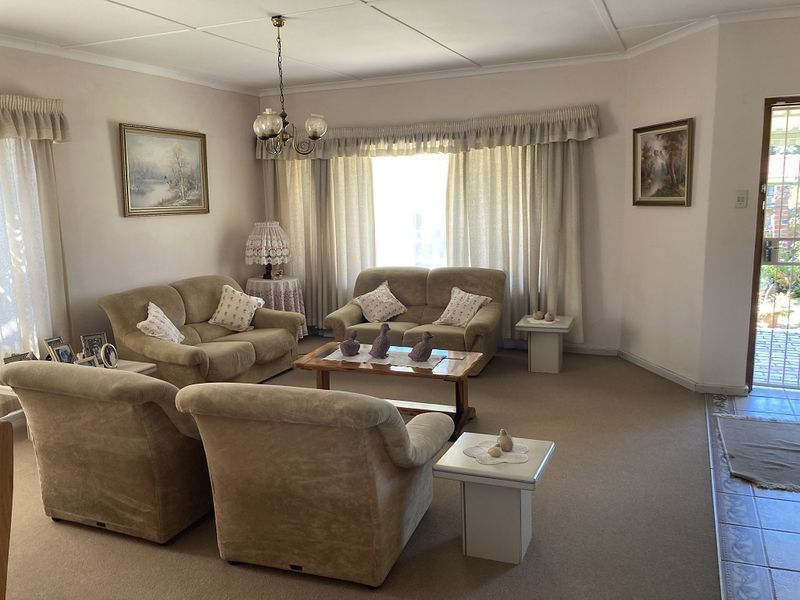 A spacious living room featuring neutral carpeting, classic ceiling tiles, and large windows with curtain treatments, providing a comfortable and traditional living space.