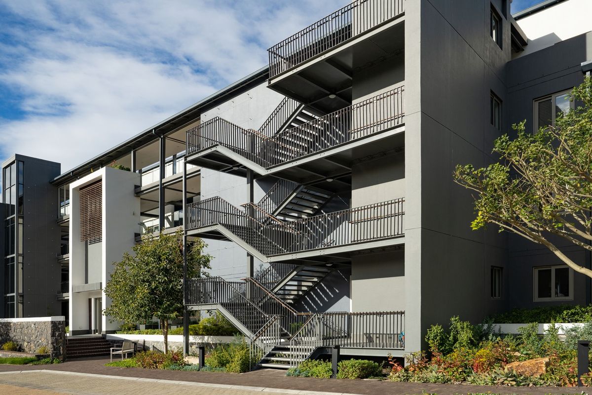 Modern apartment building exterior featuring a prominent metal fire escape staircase, private balconies, and well-maintained landscaped garden beds along a paved walkway.