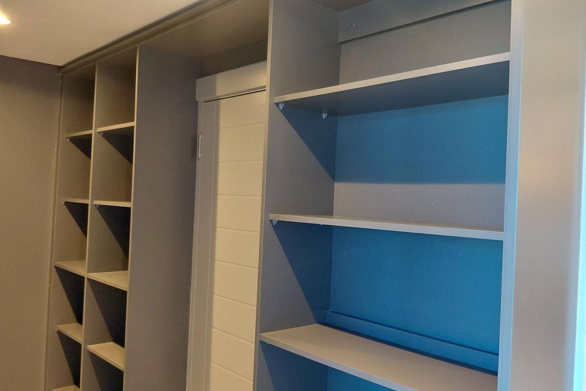 A clean bedroom interior featuring extensive built-in grey shelving units and a modern white internal door.