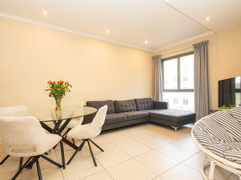 A bright open-plan living and dining area featuring neutral tiled flooring, recessed downlighting, and large windows allowing for ample natural light.