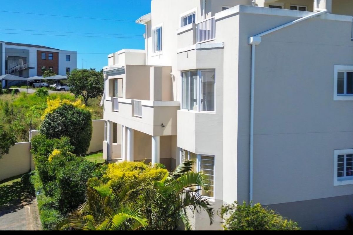 A view of the rear elevation of the apartment building, featuring balconies, white exterior walls, and landscaped garden surroundings.