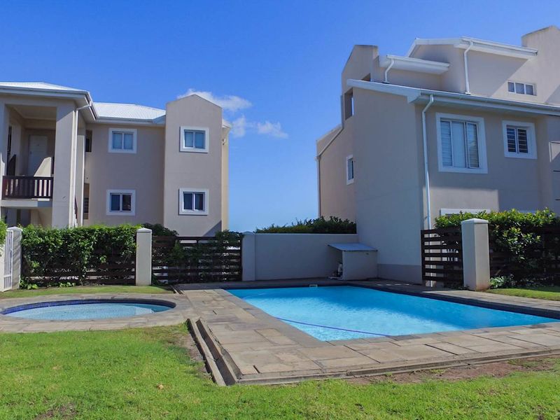 A well-maintained communal pool and spa area featuring paved decking, surrounded by landscaped lawns and timber privacy fencing between apartment buildings.