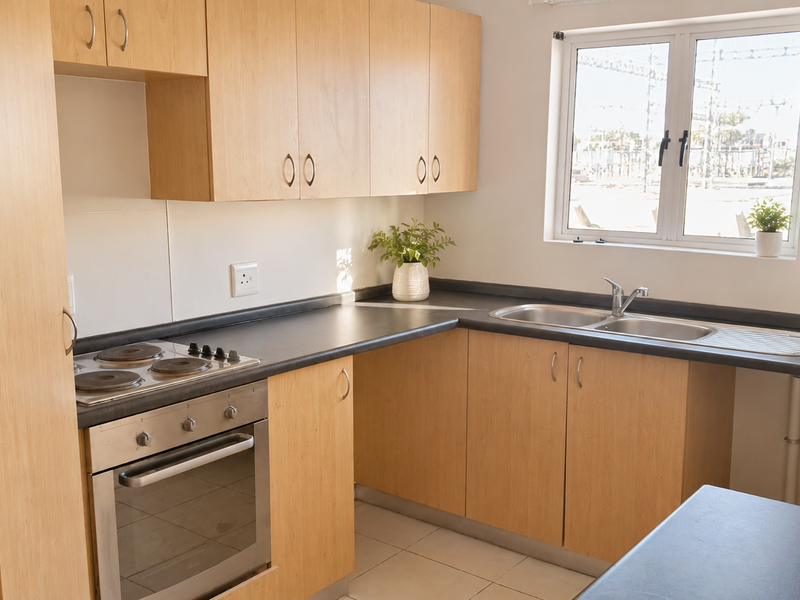 A functional kitchen featuring light wood cabinetry, dark laminate countertops, a built-in electric oven and hob, and a stainless steel double sink.