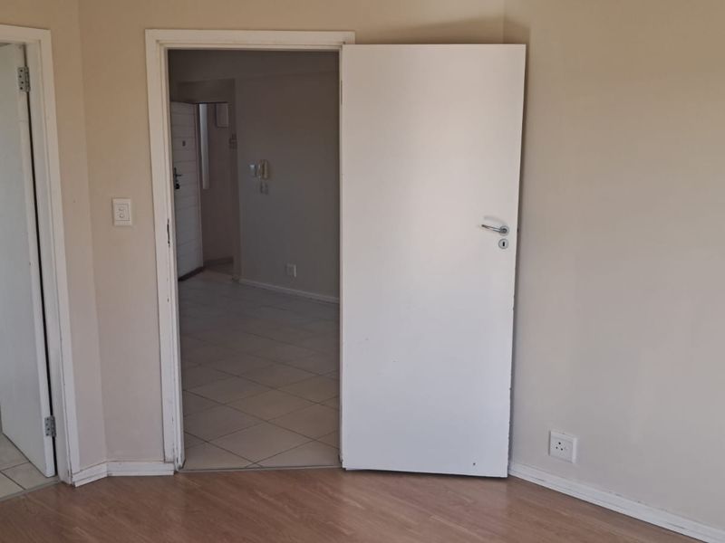 A view of an empty hallway connecting to a bathroom and adjacent living area, featuring neutral wall finishes and a mix of tiled and laminate flooring.
