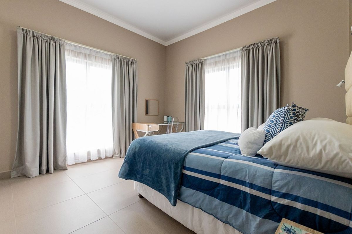 A well-lit bedroom featuring neutral wall tones, tiled flooring, and dual windows with curtain treatments, providing a bright and comfortable living space.