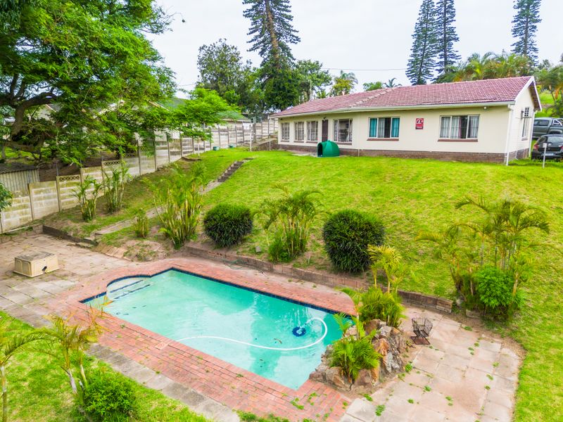 Rear exterior view featuring a swimming pool with brick coping, a paved patio area, and a large sloped lawn leading to the main house.