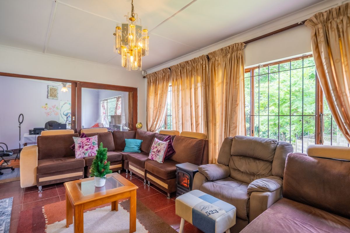 A spacious living room featuring tiled flooring, a decorative crystal chandelier, and large windows with security bars, offering a view of the garden.