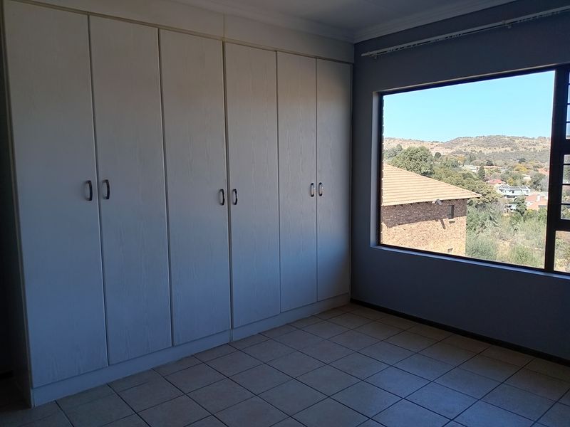A bright, empty bedroom featuring built-in cupboards, tiled flooring, and a large window offering scenic views of the surrounding landscape.