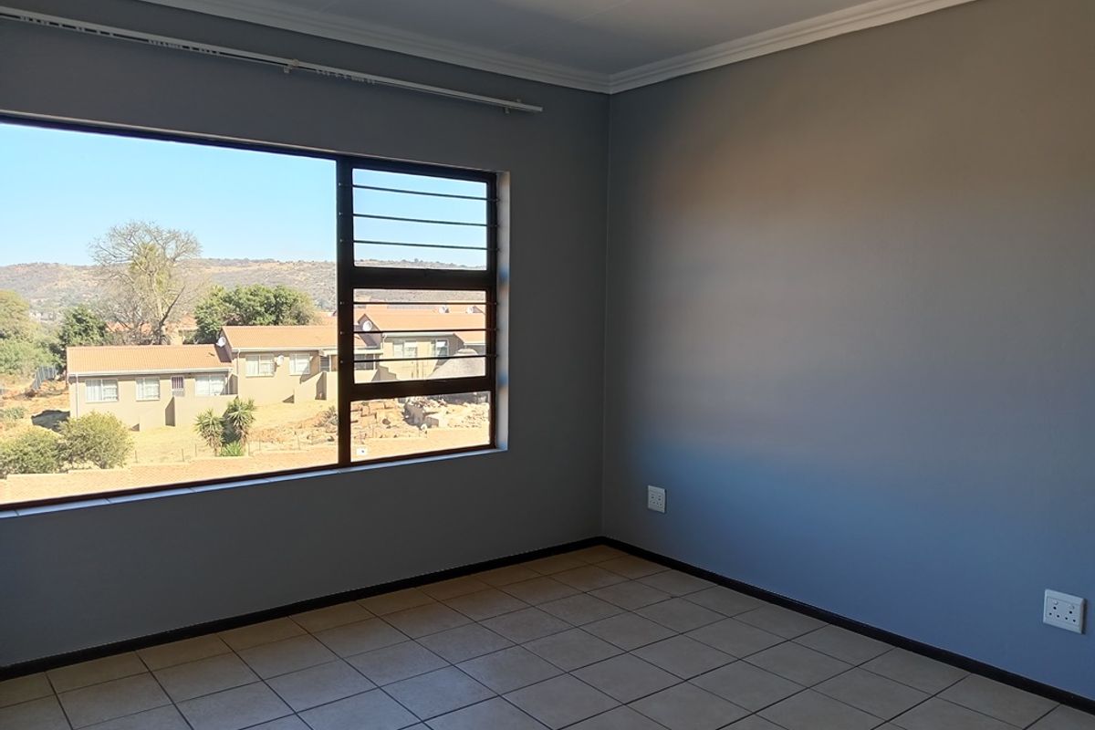 A bright, empty bedroom featuring tiled flooring, neutral grey walls, and a large window with security bars offering a suburban view.
