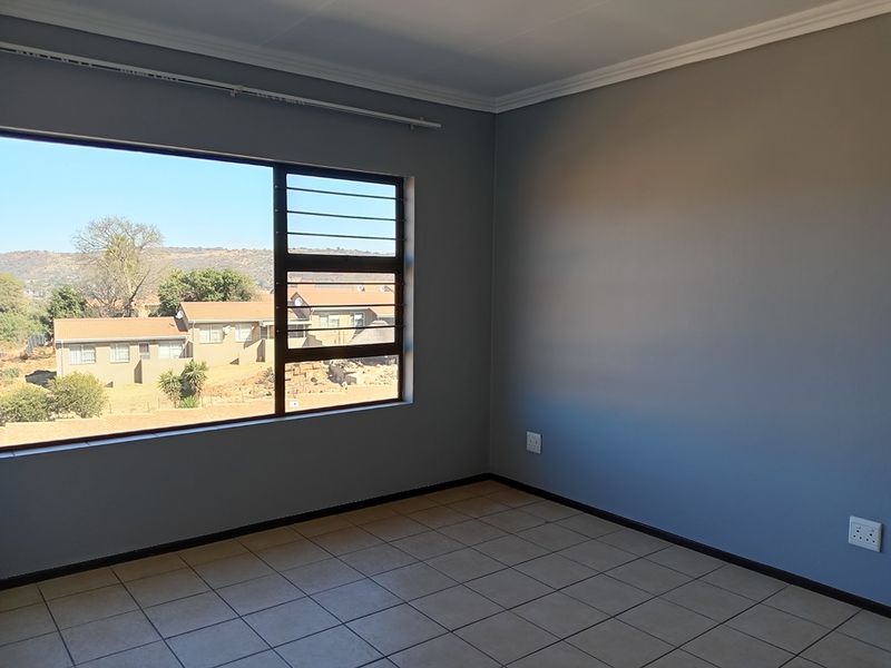 A bright, empty bedroom featuring tiled flooring, neutral grey walls, and a large window with security bars offering a suburban view.