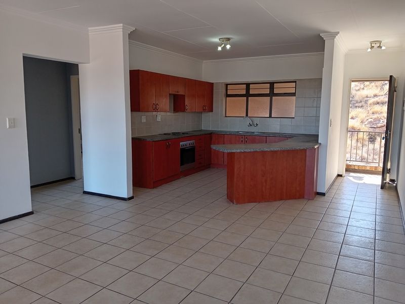 Open-plan kitchen featuring wooden cabinetry, granite countertops, and integrated appliances, flowing into a spacious tiled living area with direct access to an outdoor balcony.
