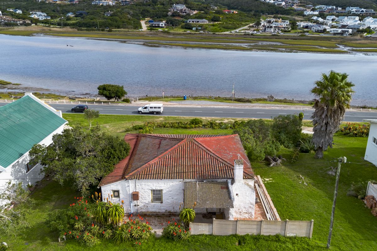 Aerial view of a coastal property featuring a traditional house with a tiled roof, covered patio, and garden, situated near a lagoon and road.