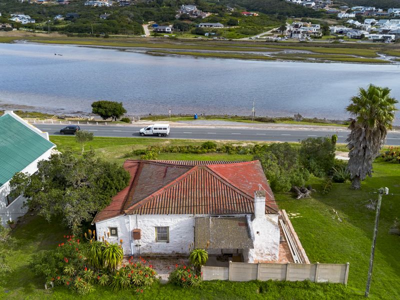 Aerial view of a coastal property featuring a traditional house with a tiled roof, covered patio, and garden, situated near a lagoon and road.