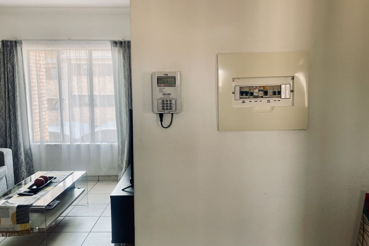 A living room area featuring a wall-mounted prepaid electricity meter and a distribution board, with tiled flooring and natural light from the window.