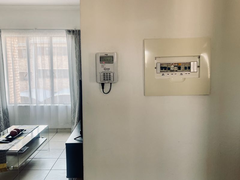 A living room area featuring a wall-mounted prepaid electricity meter and a distribution board, with tiled flooring and natural light from the window.