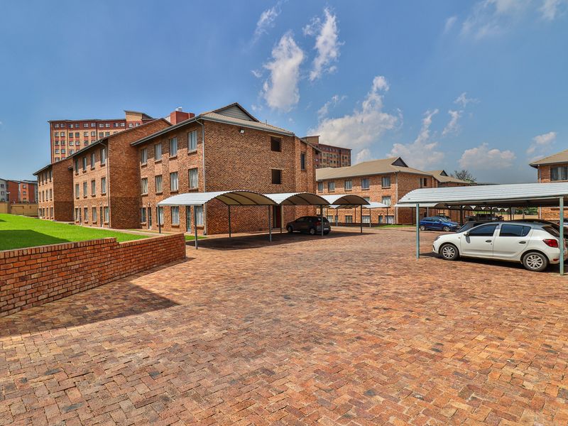 Exterior view of the apartment complex featuring brick facade buildings, paved parking area with covered carports, and a well-maintained lawn.