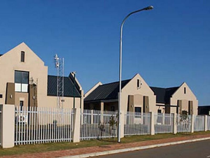 A street-level view of a residential complex featuring modern architectural design with metal roofing, stucco facades, and secure steel picket fencing.