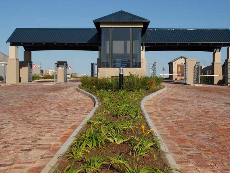 A well-maintained secure estate entrance featuring a modern gatehouse, boom gates, and a brick-paved driveway with a landscaped central island.