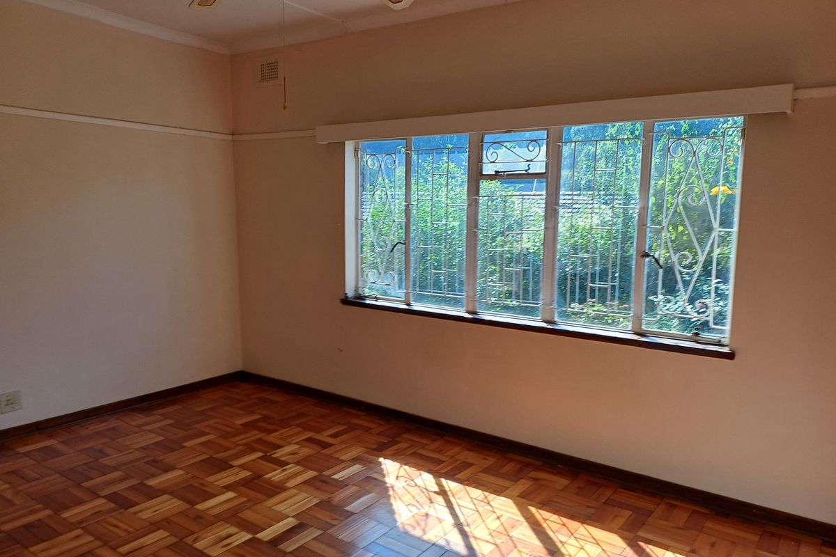 A bright, empty bedroom featuring classic parquet flooring, a ceiling fan, and large windows fitted with security bars.