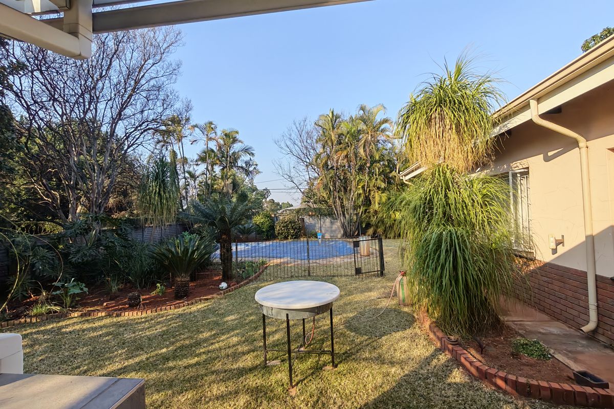 A rear garden view featuring a swimming pool with a safety fence, established landscaping, and a covered patio area.