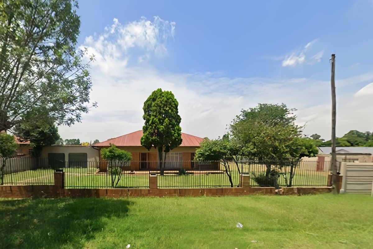 Front view of a single-story residential house featuring a metal roof, established garden, and secure perimeter fencing with brick pillars.