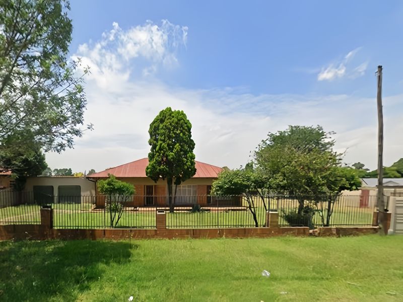 Front view of a single-story residential house featuring a metal roof, established garden, and secure perimeter fencing with brick pillars.
