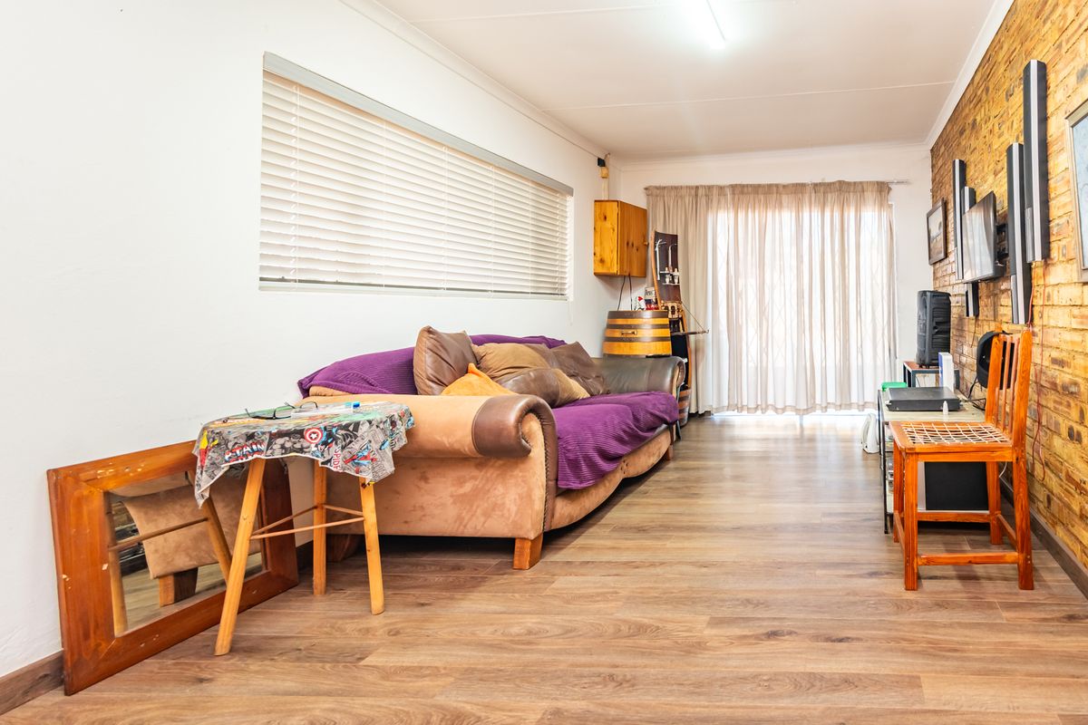 A living room featuring laminate flooring, an exposed brick feature wall, and natural light through venetian blinds and a sliding glass door.