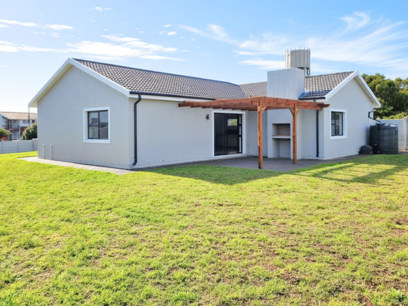 Rear view of a modern house featuring a spacious lawn, a built-in braai area, a wooden pergola, and a water tank.