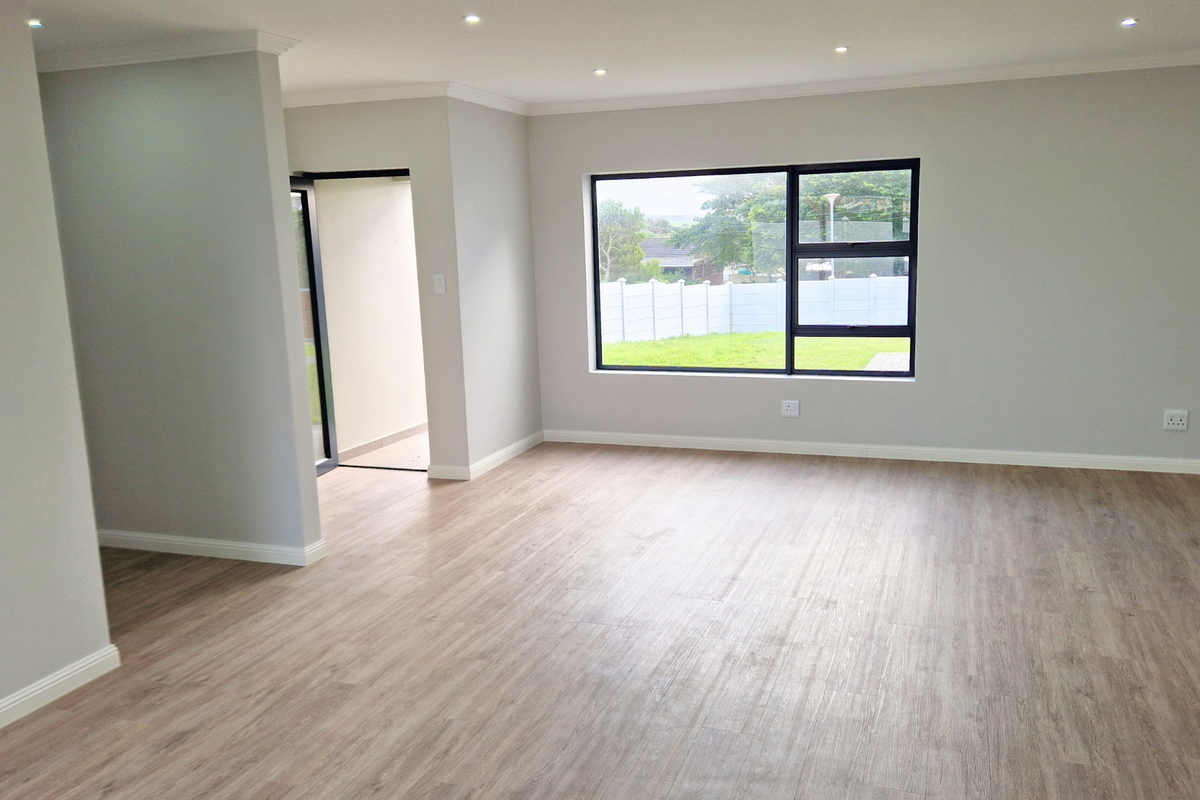 A bright, spacious, and empty living area featuring modern laminate flooring, recessed downlights, and large aluminium-framed windows with a garden view.