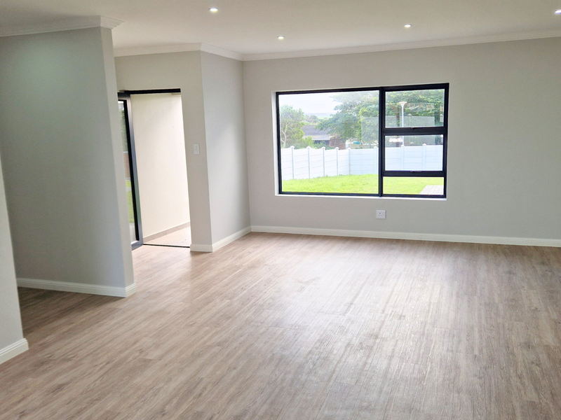 A bright, spacious, and empty living area featuring modern laminate flooring, recessed downlights, and large aluminium-framed windows with a garden view.