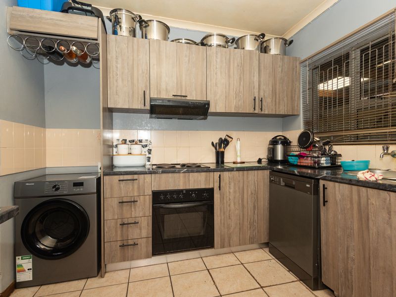 A compact kitchen featuring wood-look cabinetry, dark laminate countertops, built-in oven and hob, and integrated laundry space with tiled flooring.